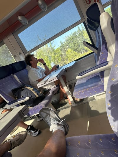 The image shows the interior of a train carriage with two people. In the foreground, the legs and feet of two individuals are visible, one wearing grey athletic shoes and white socks, and the other wearing black and silver athletic shoes. They appear to be seated. In the middle ground, a man with curly brown hair is seated, wearing a white t-shirt and shorts. He is leaning forward with his right hand near his mouth, and he is holding a phone in his left hand. He is seated next to a folded tray table. A backpack is on the seat next to him. The train carriage has purple patterned seats and large windows. Outside the windows, trees and a bright blue sky are visible. The location is identified as Salses-le-Château, France.