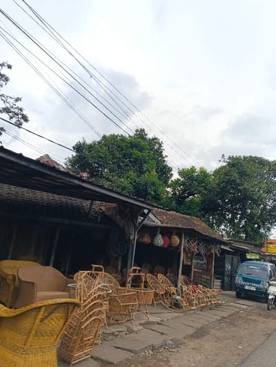 A roadside shop displays an array of rattan furniture, including chairs, sofas, and decorative items. Baskets hang from the shop's covered awning. A teal van and a white scooter sit parked on the street in front of the shop. Overhanging trees and utility wires fill the sky above the scene, under an overcast sky.