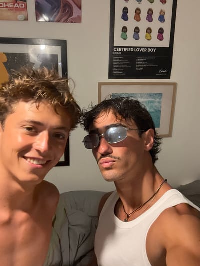 This image depicts two young adult males positioned closely together, appearing to be an indoor selfie, located in Bondi, Australia. The male on the left, partially shirtless, has light brown, messy hair and is smiling broadly at the camera. The male on the right wears a white ribbed tank top, dark wavy hair, a mustache, and reflective rectangular sunglasses, looking towards the camera with a pouting expression; a dark necklace with a small pendant is visible around his neck. The background features a light-colored wall adorned with multiple art pieces, including an unframed "RADIOHEAD the bends" poster, an abstract art print, a black-framed "DRAKE CERTIFIED LOVER BOY" poster with multiple stylized pregnant figures, and a light-wood framed picture of an ocean wave. A crumpled grey fabric, possibly bedding, is visible in the lower background. The lighting highlights features on both individuals' skin.