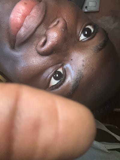 A close-up, rotated image captures a dark-skinned individual's face, with a blurred finger obstructing the lower portion of the frame. The specific location within the city of Iriebe, Nigeria, cannot be determined from the visual evidence.

**Subject:** The central figure is an individual with dark skin. Both dark brown eyes are open, fixated directly forward, with visible reflections in the pupils. The nose features prominent nostrils and a visible bridge. The upper lip, appearing full and pinkish-red, is slightly elevated, with the tongue extended and resting against it. The skin on the face exhibits a slight sheen. Dark, short hair is visible on the head.

**Objects/Foreground:** A blurred, out-of-focus finger or thumb, consistent with a human digit, extends across the lower-center of the image, partially obscuring the chin and lower facial area of the subject. The finger's skin tone appears somewhat lighter than the subject's face, possibly due to lighting or reflection.

**Environment:** The background is largely dark and indistinct. In the upper-right corner, a light-colored rectangular object with a dark switch or button, consistent with an electrical wall switch or outlet, is visible against a light brown or beige surface. The immediate environment is an indoor setting.