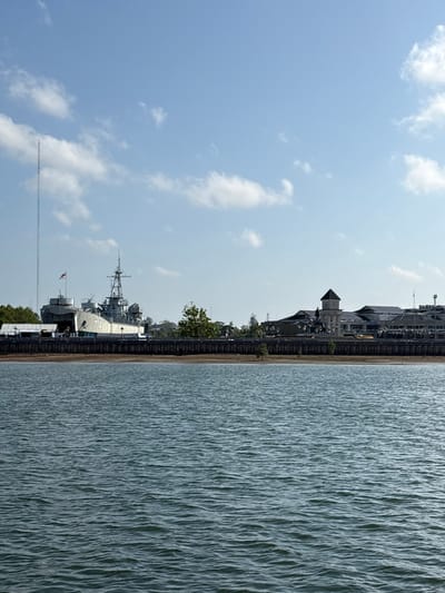 The image displays a panoramic view of the Krabi River waterfront in Krabi Town, Thailand, under a clear blue sky with scattered white cumulus clouds. The foreground consists of a body of water exhibiting minor ripples. A narrow strip of exposed mudflat or sand separates the water from a dark, elevated concrete embankment or quay wall that runs horizontally across the midground.
On the left side of the embankment, the grey-painted Royal Thai Navy warship, HTMS Lanta (hull number 714), is prominently docked, functioning as a museum. The vessel features a multi-tiered mast with radar equipment, gun turrets, and the Thai flag flying from its stern. A tall, slender antenna tower rises vertically to the left of the ship. Green foliage from trees is visible around and behind the warship.
To the right, a series of modern buildings with dark roofs are situated along the waterfront. A distinctive white building with a prominent dark-roofed architectural tower, characteristic of the Krabi Marine Esplanade, stands out among these structures. Additional buildings and trees extend further to the right. The scene appears devoid of discernible human activity or specific interactions.