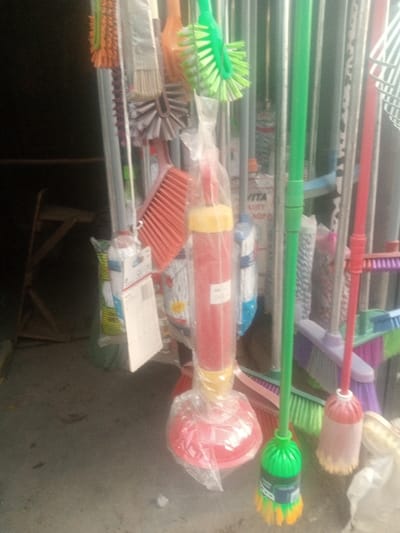 The image displays a retail arrangement of household cleaning implements in Kofar Ruwa, Nigeria. A red and yellow toilet plunger, encased in clear plastic packaging, occupies the central foreground. To its left, various brushes with orange, grey, and red bristles are suspended, along with a white product package featuring a barcode. Immediately to the right of the plunger, a green-handled broom with vibrant green and yellow bristles is prominent. Further right, another broom with a red handle and light-colored bristles is visible. The background reveals additional cleaning tools, including mops and brushes with purple, pink, and green components, with some packaging bearing the partial text "VITA QUALITY PRODUCTS." A dark, blurred area on the left side of the frame shows a partial view of a wooden stool or stand. The floor is a light grey concrete surface. No individuals are present in the scene.