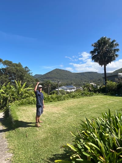 The image depicts a man standing on a grassy lawn, pointing upwards with his right hand towards a tall palm tree. In the background, a mountain range covered in dense vegetation is visible, with a few houses scattered along the lower slopes. The sky is blue with scattered white clouds. The man is wearing a dark blue shirt, shorts, hiking boots, and a white cap. The scene appears to be in Stanwell Park, Australia, based on the characteristic landscape and vegetation.