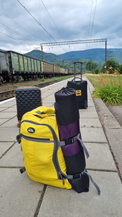 The image depicts a train platform in Kostryna, Ukraine, on an overcast day. Several pieces of luggage are arranged on the concrete platform. In the foreground, a large yellow backpack is prominent, with a rolled-up dark sleeping bag or mat strapped vertically to its front. To the left of the yellow backpack, a small, black, geometric-patterned suitcase with brown trim stands upright. Behind these, a larger black rolling suitcase with a visible handle is positioned further down the platform. In the background, a long train of freight cars sits on parallel tracks, and rolling, forested hills are visible under a cloudy sky. Orange flowers bloom along the edge of the platform to the right. The scene suggests a traveler or travelers are waiting for or have just arrived by train.