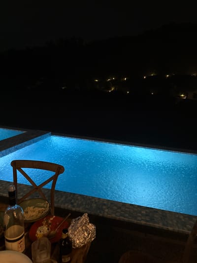 The image, taken at night, shows a scene adjacent to an illuminated pool, likely in Jinhua, China, judging by the style of the architecture and pool design common in the region. The pool is rectangular and brightly lit with blue light, reflecting off the rippled surface. To the right of the pool, there's a table littered with remnants of a meal, including plates with leftover food, aluminum foil, and bottles. A wooden chair is situated next to the table. In the background, sparse lights suggest either a residential area or landscaping. The atmosphere seems relaxed, implying a casual gathering or meal.