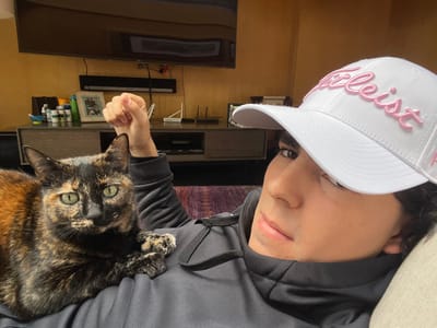 The image depicts an indoor scene located in Fraccionamiento y Club de Golf los Encinos, Mexico. A male individual, appearing to be lying down, is positioned in the foreground, with a tortoiseshell cat resting on his chest or lap. The individual wears a dark-colored hooded sweatshirt and a white baseball cap featuring "Titleist" embroidered in pink script. His right hand is clenched into a fist and positioned behind the cat's head. The cat, exhibiting a distinct black, orange, and brown fur pattern with prominent green eyes, is looking directly at the viewer. The background reveals a residential setting with a large, dark-colored flat-screen television mounted on a light brown or mustard-yellow wall. Beneath the television is a dark media console, upon which various objects are arranged: a collection of small bottles or containers, a framed picture, and multiple electronic devices, possibly routers, characterized by visible antennas. Wiring is discernible connecting the devices below the television. A light-colored, textured cushion or fabric is partially visible in the lower right, and a dark, patterned fabric section is present on the left side of the frame.