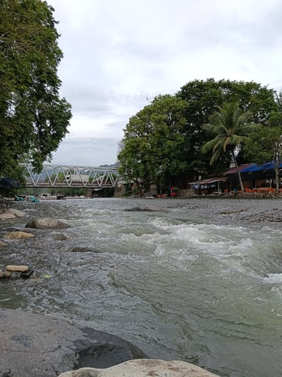This image depicts a wide, fast-flowing river with rocky banks. In the background, a white metal truss bridge spans the river, with a truck visible on it. On the right bank of the river, there are several small buildings with blue and red roofs, appearing to be shops or food stalls, with tables and chairs set up outdoors. Lush green trees, including a prominent palm tree, line the riverbank. The sky is overcast. On the left bank, more trees are visible, framing the scene. Some scattered rocks are in the foreground on the riverbed. The overall impression is a scenic natural environment with some human infrastructure. The location is identified as Meurah, Indonesia.