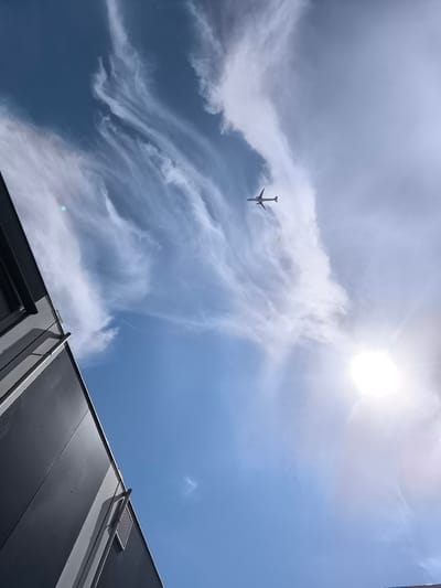 An airplane is captured in silhouette against a bright, partly cloudy sky. The plane is positioned in the upper right quadrant of the image, flying away from the viewer. Wispy cirrus clouds streak across the sky, creating a dynamic pattern. The sun is a bright, blown-out disc in the upper right corner, its rays fanning out. In the lower left portion of the image, the corner of a modern, dark gray building is visible, angled upwards towards the sky. The building has a segmented facade and a prominent horizontal conduit or pipe running along its side, with a louvered vent below it. The scene appears to be an outdoor setting, likely near an airport or an area with regular air traffic, given the presence of the airplane and the open sky. The location is Uithoorn, Netherlands.