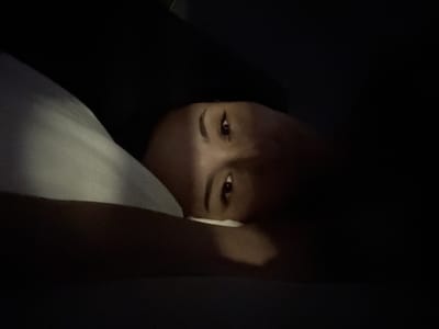 The image shows a close-up of a person's face as they lie in bed, partially obscured by darkness. Their eyes are open and looking towards the camera, with a faint light reflecting in them. The person's head is resting on a pillow, which is visible on the left side of the frame. The overall impression is one of intimacy and quiet, perhaps taken at night or in a dimly lit room. The location in Doma 1096 Km, Russia, suggests a domestic setting.