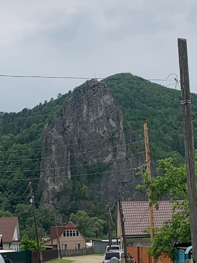 The image depicts a mountainous landscape with a prominent rock face in the background, likely a prominent landmark in Kavalerovo, Russia. The rock face is steep and appears to be covered in vegetation on its slopes. In the foreground, there are signs of human habitation, including houses with reddish-brown tiled roofs and wooden utility poles with visible electrical wires. Several vehicles, including a white SUV, are parked on a road or pathway. The overall scene suggests a rural or semi-rural setting within or on the outskirts of the city. The sky is overcast, indicating a cloudy day.