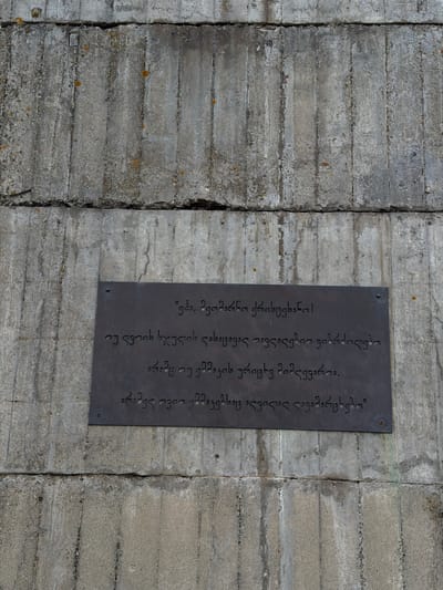 A landscape, eye-level, full shot photograph depicts a grey concrete wall with a dark grey metal sign affixed to it. The wall is made of stacked, horizontal concrete blocks with vertical seams between them. There are slight variations in texture and tone across the blocks, adding a sense of age and weathering.
The sign is rectangular and features text in Georgian script. The text is engraved or etched into the metal, creating a contrast between the dark metal and the lighter characters. The sign appears to be attached to the wall with screws or bolts, visible at the corners.
The image is clean and well-lit, capturing the texture of both the concrete and the metal sign. The location of the sign is in the city of Zemo Nichbisi, Georgia.