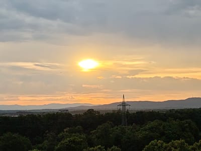 The image captures a panoramic view of Freiburg im Breisgau, Germany, at sunset. The sun, a bright orb, is positioned slightly left of the center in the upper half of the frame, casting a warm golden glow across the sky. Wispy clouds, tinged with orange and grey, stretch across the horizon. In the middle ground, a dense, dark green forest forms a lush canopy. Prominently situated within this forest is a tall, lattice-like electricity pylon. Beyond the trees and pylon, a series of layered, dusky blue hills recede into the distance. The overall scene is serene, with the natural landscape dominating the frame, punctuated by the man-made structure of the pylon.