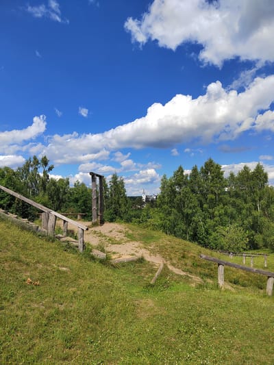 A wide-angle shot captures a sunny day with a bright blue sky filled with fluffy white clouds. In the foreground, a grassy hill slopes down, with a worn wooden railing guiding a path. Further up the hill, a sandy area features a rustic wooden structure resembling a gallows. Behind this, a dense line of green trees forms the backdrop, and in the distance, a white steeple of a church is visible against the horizon. The overall setting appears to be a rural or historical park area.