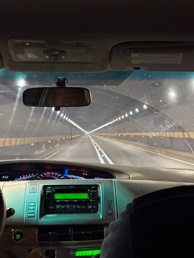 The image, taken from inside a moving vehicle, depicts the interior of the car and the view through the windshield of a long tunnel illuminated with rows of lights. The car's dashboard is visible on the right, displaying the radio tuned to CHI 104.5MHz and a temperature of 28 degrees Celsius. The rearview mirror is in the center of the frame. The environment outside the car is a long tunnel with repeating lights, indicating a consistent, linear path. Without further details, the specific location in Catia La Mar, Venezuela, cannot be determined.