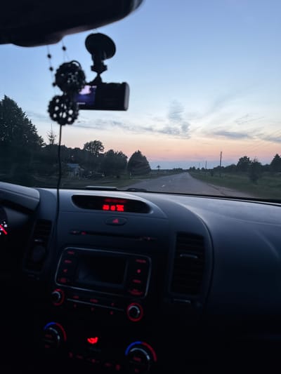 The image shows the interior of a car looking out at a road at dusk. The dashboard of the car is visible in the foreground, with a red digital display showing "20:18". A dashcam is mounted on the windshield. The sky is a gradient of blue and pink, with some clouds. Trees and houses are visible on either side of the road. The location in the city of Petrovskii, Russia cannot be determined from the image.