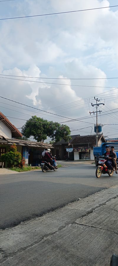 In Sukarame, Indonesia, a street scene unfolds under a partly cloudy sky. Two motorcycles are visible on the asphalt road. The first motorcycle, traveling from right to left, carries three individuals: two young males in the front and rear, and a male in the middle, all wearing casual clothing. The second motorcycle, positioned further to the left and moving towards the foreground, has two occupants wearing helmets. One rider wears a red jacket, and the passenger has a gray head covering.
The environment includes a blue truck parked to the right of the road, adjacent to buildings. These buildings appear to be shops or residences, with one featuring a sign that reads "DEVO AIR MINUM." A large tree with dense green foliage stands between two of these structures. Utility poles with numerous wires are prominent in the background, indicating the presence of electrical infrastructure. The road itself shows signs of wear, with a cracked concrete edge on the right side. The lighting suggests it is daytime, possibly late afternoon, given the shadows cast.