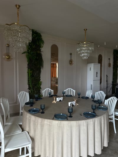The image captures an indoor dining setup. A long, draped table occupies the central space, set with plates, glasses, and silverware. White chairs are arranged around the table. Above, ornate chandeliers hang from the ceiling. Decorations of greenery adorn the walls. A tall, white appliance stands against the wall in the background. The scene appears to be in Marneuli, Georgia, based on visible text on a sign.