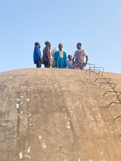 The image depicts six individuals standing on the apex of a large, weathered, concrete or stucco dome structure in Barako, Nigeria. The dome's surface is light brown/grey and shows extensive cracking and areas of chipped material. On the right side of the dome, a series of metal rungs, forming a ladder, extends downwards.
The six individuals, presumably adult males or adolescents, are silhouetted against a clear, light blue sky. From left to right: one male wears a blue long-sleeved shirt and dark trousers, looking towards the center. Next is a male in a striped shirt and a light-colored cap, also looking towards the center. The central figure wears a vibrant blue traditional robe with gold embroidery and a light cap, facing forward and smiling. Partially obscured behind and to the right of him is a smaller individual in a light blue garment and cap. To the right of the central figure, a male wears a light-colored short-sleeved shirt, a grey vest, and dark trousers, facing forward and smiling. Behind the central group, two light-colored, spherical or urn-shaped objects, possibly decorative elements or components of the structure, are visible on the dome. The individuals appear to be gathered on the dome, with some looking towards the camera.