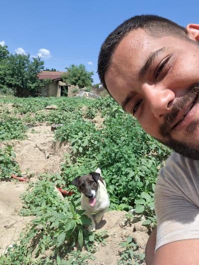 A man with dark hair and a beard is taking a selfie with a dog in a rural setting. The man's face is close to the camera, with sweat visible on his forehead. The dog, a Jack Russell Terrier mix with black and white markings, stands in front of him, panting with its tongue out. The background shows a field of green plants, a dirt path, and a dilapidated building with a red roof. The sky is clear and blue, with a few white clouds. This scene is located in the village of Dolna Malina, Bulgaria, a location known for its agricultural lands and rustic charm.