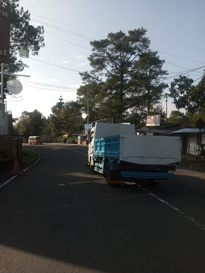 The image captures a road scene on a sunny day, possibly in the Phouviang-Gnai area of Laos, though the exact location cannot be confirmed from the visual information alone. The focus is on the back of a blue and white flatbed truck driving along the road. The road appears to be asphalt and has a white line marking the lane. The truck has "MEKANIK" and "TOBRUT" visible on the lower rear section. On the left side of the road, there is a sign with "ATM" visible on top. There are trees along the roadside, and in the background, a yellow van is seen driving in the opposite direction. There is a structure with stalls or shops on the right side of the road, along with a billboard in the distance. Utility poles and wires are visible overhead.