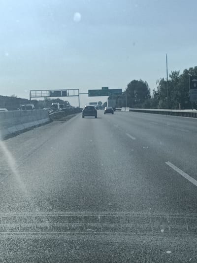 A car is driving on a highway. The car is dark in color and is in the right lane. The highway has multiple lanes and is surrounded by trees and buildings. There are also overhead signs indicating directions. The sky is blue and there are a few clouds. The image appears to be taken during the daytime. The location is Kunice, Czechia.