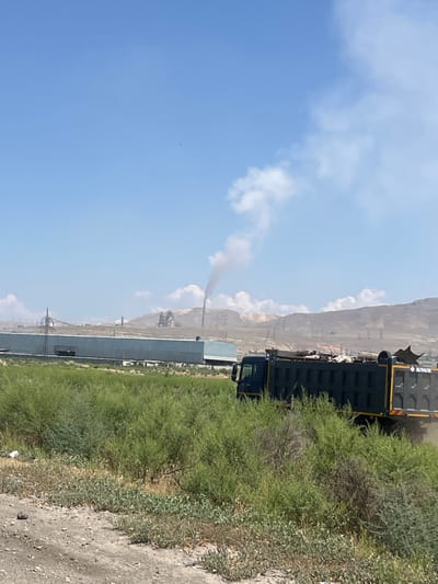 This vertical media file depicts an outdoor scene in Surenavan, Armenia. The foreground is dominated by lush green vegetation on the left and a dusty, gravelly path on the right. In the midground, a large, dark-colored truck is visible, its bed filled with what appears to be white material. The truck has yellow accents and visible text that reads "SHAVK" in white lettering on its rear.

In the background, a long, industrial-looking building with a grey facade stretches across the frame. Beyond the building, a mountainous landscape rises under a bright blue sky dotted with white clouds. A plume of dark smoke is rising from what appears to be a smokestack in the distance, suggesting industrial activity. The lighting indicates it is daytime, likely midday or afternoon, given the brightness of the sky. The overall impression is of a semi-arid, industrial or rural environment. No people are clearly visible in the scene. The image seems to capture a moment of the truck's activity or presence in this location.