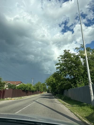 A street scene in Muzychi, Ukraine, on a day with dramatic cloud formations. The sky is a mix of bright blue patches and dark, heavy storm clouds, particularly on the left side of the image. A tall, grey utility pole stands prominently on the right side of the road, with sunlight glinting off its surface. Dense green trees line the right side of the road, with their branches casting shadows on the asphalt. On the left side of the road, a reddish-brown corrugated metal fence runs along the property line of houses, which are partially visible behind the fence and trees. The road itself is paved and has a white line marking the center. The perspective suggests the photo was taken from a vehicle. The environment appears to be a residential or semi-rural area. There are no people or animals clearly visible in the scene. The overall atmosphere is dynamic due to the contrasting sky conditions.