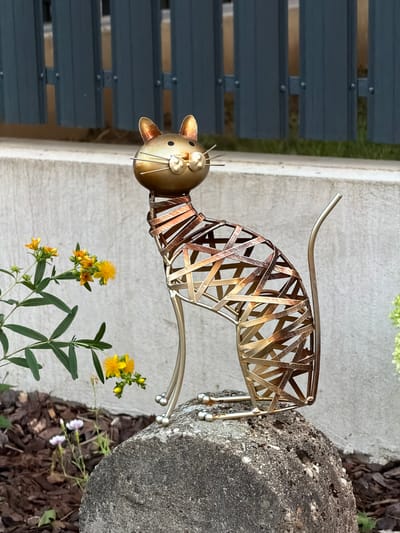 The image shows a metallic garden sculpture of a cat, crafted from interwoven bands of gold and copper-toned metal. The cat sculpture is positioned on a rough-textured, grey rock. To the left of the sculpture, a sprig of small yellow flowers with green leaves is visible. In the background, a grey concrete wall and a dark grey slatted fence are present. The ground beneath the sculpture and to the left is covered with dark mulch, with a few small white and pink flowers also visible in the mulch. The scene appears to be an outdoor setting, likely a garden. The provided information that the image is from Tiszaszentmárton, Hungary, cannot be visually confirmed from the image itself.