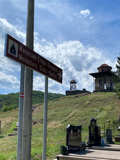 The image captures a view of the Monastery of Stara Pavlica, located in the Raska Municipality of Serbia. In the foreground, a directional signpost points to the right, indicating "Monastery Stara Pavlica" in both Serbian and English. The sign features an icon of a church. Several metal poles support the sign, and a small red and white marker is attached to one of them. To the right of the sign, a graveyard with several black headstones is visible.

In the mid-ground, a grassy hill rises, with a path of stone steps ascending its slope. Further up the hill, the ruins of the Monastery of Stara Pavlica are situated. The main structure is a stone building with a domed roof and arched windows. Adjacent to it is a wooden bell tower. The background shows a lush green, tree-covered hillside under a bright blue sky with scattered white clouds. The overall scene depicts a historical and religious site in a natural setting.
