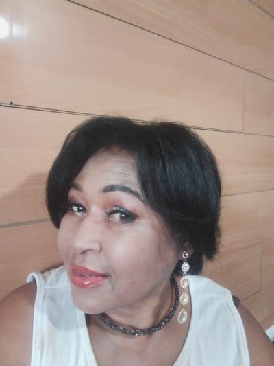 A medium shot selfie shows a Black woman with dark, layered hair, wearing a white sleeveless top, a sparkling dark choker necklace, and a dangling earring with multiple clear stones. She has on makeup, including red lipstick and eyeshadow. The background is a wood-paneled wall. The image provides no information about the city of Kuntuu, Kyrgyzstan.