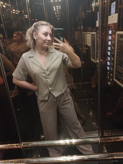 A woman stands in an elevator, taking a selfie in a mirrored wall. She is wearing a light brown button-up shirt and matching pants. Her blonde hair is pulled up in a ponytail. The elevator walls are reflective, showing multiple reflections of the woman and the elevator interior. The elevator control panel with illuminated buttons is visible on the right side of the frame. The overall scene suggests an urban indoor environment, possibly a hotel or commercial building.