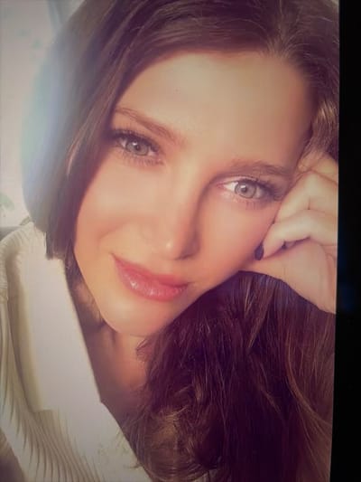 The image displays a brightly lit, close-up portrait of an adult female. Her head is tilted slightly, and she exhibits a subtle smile. She has light-colored eyes (blue-grey) and long, dark brown hair that cascades over her right shoulder and partially obscures the right side of her face. Her left hand, with fingernails painted a dark color, rests against her right cheek and jawline. She is wearing a light-colored, possibly cream or white, collared garment with a visible ribbed texture on the left side of the frame. A notable lens flare is present in the upper left quadrant of the image. The background is unfocused and indistinct, offering no discernible environmental features. The specific location within Kiruna, Sweden, is not discernible from the image content.
