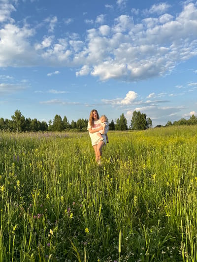 A young woman is standing in a field of tall grass and wildflowers. She is holding a baby in her arms. The woman is wearing a white t-shirt and white shorts. The baby is wearing a white and grey patterned outfit. The field is full of yellow and purple flowers. In the background, there is a line of trees. The sky is blue with white clouds. The image appears to have been taken in the afternoon, with the sun casting a warm glow on the scene. The location is the village of Koloksha, Vladimir Oblast, Russia.