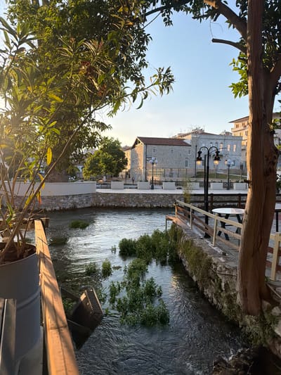 The image presents a view of the city of Drama in Greece. The scene is framed by trees and their foliage in the foreground, partially obscuring the overall view. A river runs through the center of the frame, reflecting the ambient light. The river banks are lined with stone walls and vegetation. Beyond the river, buildings with light-colored facades and traditional architectural elements are visible. Street lamps are positioned along the river and near the buildings, indicating urban development. Tables and chairs can be seen suggesting a restaurant or cafe seating area adjacent to the river. The sky is clear and bright, suggesting daylight conditions.