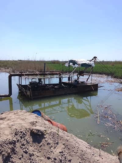 The image depicts a rusty, makeshift dredging or pumping barge situated in a murky body of water, potentially a river or lake, in Syrdarya, Uzbekistan. The barge is constructed with a metal frame and houses several dark-colored mechanical components. A makeshift canopy covered in white plastic sheeting sits atop the barge.
The water is greenish-brown in color and contains debris, including plant matter and sticks. The barge's reflection is visible in the water.
In the foreground, there is a large pile of dirt or sand with a discarded barrel partially buried within it. The landscape surrounding the water is mostly flat, with some tall grasses and vegetation in the background. The sky is clear and blue.