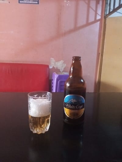 The scene is set indoors in Kisii Central ward, Kenya. A clear, faceted glass containing a golden liquid topped with approximately 30mm of white foam is positioned on the left side of a dark, reflective table surface. To its right, an amber-colored glass bottle with a "White Cap Lager" label, depicting a mountain graphic, stands upright. The bottle appears partially full. The background consists of a reddish-orange painted wall exhibiting areas of minor paint or plaster peeling. A segment of a red upholstered bench or seating is visible against the wall on the left, next to a purple plastic stool or container. On the far right, a dark, vertical structural element, possibly a door frame or grating, is partially visible. A small, illegible sign is affixed to the upper left portion of the wall. No individuals are present in the image.