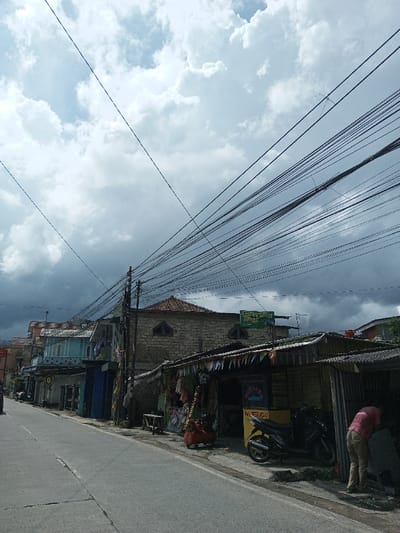 The image presents a street scene in Cibadak, Indonesia, characterized by a heavily clouded sky overhead, showing lighter cumulus clouds on the left transitioning to dense, dark nimbostratus-like clouds towards the right. A paved road occupies the lower-center of the frame, receding into the distance. Along the left side of the street, a sequence of multi-story buildings, some with balconies (one featuring a light blue railing), line the thoroughfare. On the right side, multiple dark utility poles support a dense network of black electrical and communication wires spanning across the frame. Behind these poles, a substantial building constructed from light-colored stone or brick features a tiled roof and a distinctive arched window. Above this building, a green sign with white text, likely reading "RAUDLATUL ATHFAL ISLAM," is mounted. In the foreground on the right, a series of open-fronted roadside stalls with corrugated metal roofs are visible, adorned with colorful triangular pennants and displaying various merchandise. A yellow vendor cart is positioned next to one of these stalls, with a dark-colored motorcycle parked alongside it. One individual, wearing a reddish-pink shirt and light-colored pants, is observed bending over near the right edge of the frame, attending to an object on the ground next to the motorcycle.