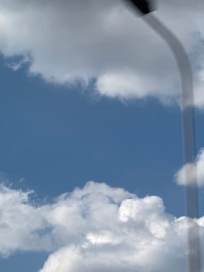 The image displays a bright blue sky partially obscured by scattered white and grey cumulus clouds. A light-colored, curved pole, consistent with a street light or utility pole, extends from the upper right corner, curving downwards towards the center. The pole's apex features a darker, indistinct object. A faint shadow runs along the length of the pole. The scene is an outdoor environment, devoid of people, specific buildings, or vehicles. No visual identifiers within the image confirm its specific location in Przyszowice, Poland.