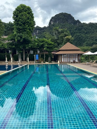 The image shows a swimming pool with turquoise tiled water and blue lane markers in Thap Put, Thailand. The pool is surrounded by a wooden deck and lounge chairs, with a thatched-roof pavilion and a building in the background. Lush green trees and a large, rocky mountain covered in vegetation dominate the landscape. The sky is overcast with dramatic clouds. No people are visible in the image.