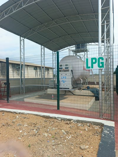The image depicts an LPG (Liquefied Petroleum Gas) filling station, possibly located in Zonkwa, Nigeria. The station is enclosed by a green wire fence and covered by a metal roof supported by steel pillars. Inside the enclosure is a large, white, horizontal LPG storage tank. A dispensing unit with a digital display and hose is connected to the tank. A green and white "LPG" sign is prominently displayed. The ground outside the fenced area is a mixture of dirt and gravel. In the background, a building with dark-stained walls is visible through the fencing. There are no people visible in the image.