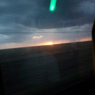The image captures a twilight scene, likely viewed from a window, given the presence of a faint green light source at the top and what appears to be a reflection or smudge on the glass. The horizon is marked by a faint line, separating a dark, blurry landscape from the sky. The sky itself transitions from a deep blue-gray at the top to lighter, softer tones near the horizon, with subtle hints of clouds. A bright, diffused light, suggestive of the setting sun, is visible low on the horizon, casting a warm glow on the lower part of the sky. The landscape below is predominantly dark and indistinct, suggesting it is either nighttime or heavily shadowed. There are no discernible people or specific structures visible in the scene. Without additional context or visual cues, it is impossible to definitively place this image within the city of Adyk, Russia.