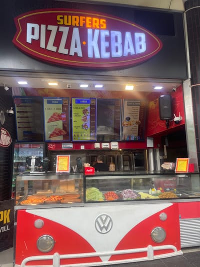 This image shows the exterior of "Surfers Pizza Kebab," a fast-food establishment located in Surfers Paradise, Australia. The facade of the shop features a prominent, brightly lit sign that reads "SURFERS PIZZA KEBAB." Below the sign, a display window showcases various food items, including pizzas, salads, and what appear to be kebab ingredients. The shop is designed with a retro, red and white color scheme, reminiscent of a Volkswagen van. There are menus displayed on the wall above the food display, detailing prices for pizza, kebab, chips, and drinks. In the foreground, the lower part of the building is decorated to resemble the front of a classic van, complete with headlights and a grille. A Coca-Cola cooler is visible to the right. Inside, a person can be seen working behind the counter, preparing food. The overall scene depicts a busy takeaway food outlet in a commercial area.
