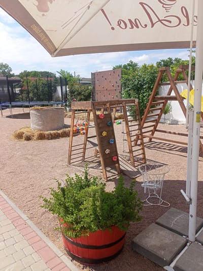 An outdoor recreational area in the city of Shabo, Ukraine, features a wooden playground structure with a climbing wall, ladder, and slide. Nearby, a large bale of hay sits on a bed of straw, with a trampoline visible in the background. A red barrel planter filled with green bushes adds a decorative element. The ground is covered in gravel, and a paved walkway lines the edge of the area. A large white umbrella provides shade, and a white metal basket-like object stands near the playground. The scene is set against a backdrop of lush green trees and a partly cloudy sky.