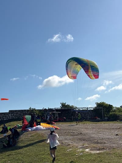 A vibrant, multicolored paraglider is suspended in a clear blue sky dotted with scattered clouds. The foreground shows a grassy area and a gathering of individuals, possibly preparing for or resting after paragliding. There are multiple sets of paragliding equipment laid out on the ground. A windsock stands in the background. The location is likely in Benoa, Indonesia, based on its known reputation as a paragliding location.