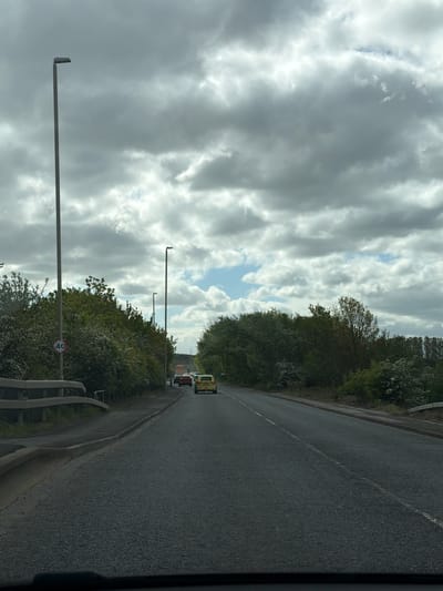 The image depicts a road in South Tyneside, United Kingdom. The road is paved and has a white line marking the center. There is a vehicle with flashing lights and other vehicles in the distance. The road is bordered by trees and bushes on both sides. There are street lights along the road. A sign indicating a speed limit of 40 is visible. The sky is cloudy with some patches of blue. The image appears to have been taken from inside a moving vehicle.