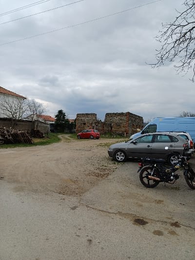 This image depicts an outdoor scene in Prdejci, North Macedonia, under an overcast sky. The foreground features a dark asphalt surface with visible oil stains, transitioning into a dirt and gravel area in the midground. On the right side of the asphalt, two black motorcycles with some reddish-brown accents are parked, one facing left, the other partially obscured behind it. A dark grey hatchback car, possibly a Seat Ibiza, is parked on the dirt/gravel surface to the left of the motorcycles, facing right. Further back, a light blue panel van is parked behind the grey hatchback.

In the left midground, a stack of wooden pallets is visible next to a low wall. Behind this, a white building with an orange tiled roof is partially visible. In the background, two prominent, partially collapsed stone and brick structures, appearing as ruins, stand centrally. To their left, a red compact car is parked on a dirt path leading into the distance. Bare tree branches are visible in the top right and left portions of the frame, and power lines span across the top of the image against the grey sky. No individuals are present or engaged in any actions within the visible scene.