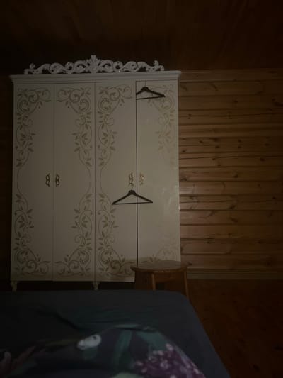 The image shows a white dresser with ornate carvings on its sides and a floral scroll pattern on its drawers. Two black hangers are placed on the top drawer. The dresser is positioned in a room with wooden paneled walls. To the right of the dresser, a portion of a dark blue or grey sofa or bed with a floral patterned throw is visible. The scene appears to be indoors, likely a bedroom, given the presence of the dresser and the sofa/bed. The lighting is dim, suggesting it might be evening or nighttime. There is no visible text in the image. The location context provided is Новая Отрадовка, Russia. The overall impression is one of a domestic setting.