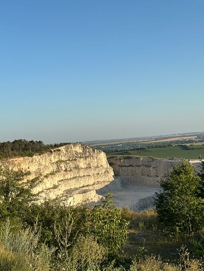 An eye-level, wide-angle shot captures a limestone quarry in Verbka, Ukraine, under a clear blue sky. The quarry, with its distinct layered rock formations, dominates the left and central portions of the frame. To the left of the quarry, a dense line of pine trees marks the edge of the landscape. In the background, a sprawling rural scene unfolds, featuring green fields, scattered houses, and distant industrial structures. The foreground is filled with lush vegetation, including grass, bushes, and trees, their leaves catching the sunlight. The overall impression is one of a vast, open landscape at dusk, with the warm glow of the setting sun illuminating the quarry walls.