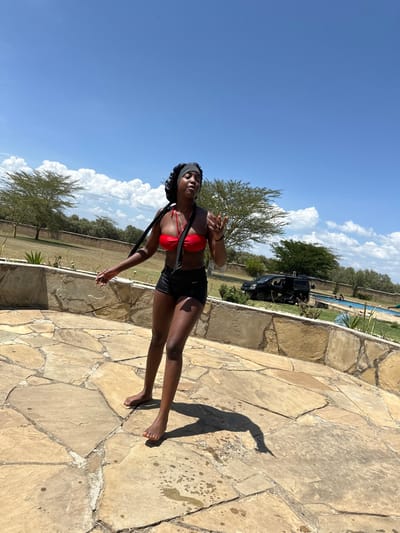 The image depicts an outdoor daytime scene in Olkaria ward, Kenya, under a clear blue sky with scattered white cumulus clouds. A single adult female with dark skin is centrally positioned in the foreground, standing barefoot on a light-brown flagstone patio. She is attired in a red strapless bikini top and black athletic shorts, with a black satin bonnet covering her head, from which two black fabric ties extend over her shoulders. Her eyes are closed, and her mouth is slightly open. Her right arm is raised with the hand slightly open, and her left arm is extended slightly downwards and to her side. Wet spots and footprints are visible on the flagstone patio around her.
In the mid-ground, a low wall constructed of rough-hewn stones separates the patio from a grassy lawn. Beyond the wall, several sparsely foliated trees, characteristic of acacia species, are present. A dark-colored utility vehicle is parked on the grassy area to the right. In the background, a blue swimming pool is visible, with two indistinct human figures partially submerged or standing by its edge. The bright sun casts a distinct, elongated shadow of the primary individual onto the flagstone surface. The overall environment suggests a recreational or leisure setting within an arid or semi-arid landscape.