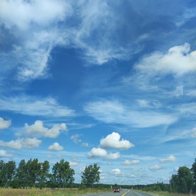 The image depicts a highway scene with a red car driving on the right lane. The road stretches into the distance under a vast blue sky filled with scattered white clouds. On the left side of the road, a line of trees stands in a field, and on the right, more trees and vegetation line the roadside. The location is identified as Marfino, Russia.